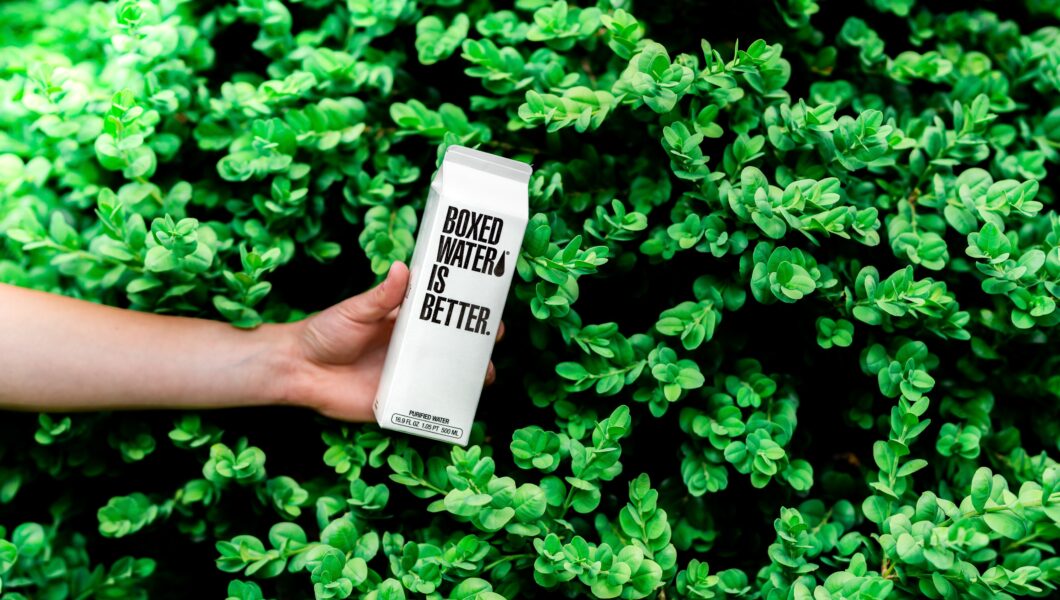 tło z parkanu, ręka trzyma biały karton z napisem boxed water is better);