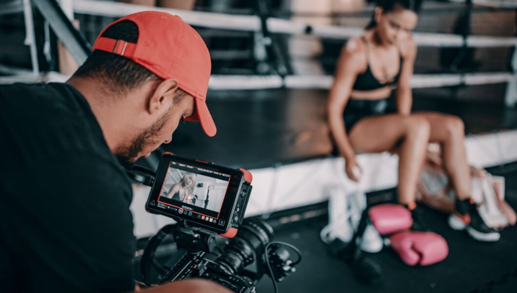 mężczyzna z brodą w czerwonej czapce stoi tyłem, jest za aparatem foto, robi zdjęcie wysportowanej dziewczynie);